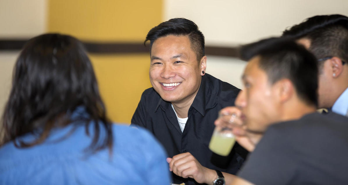 Photo of person eating at a table smiling while talking to folks seated next to them
