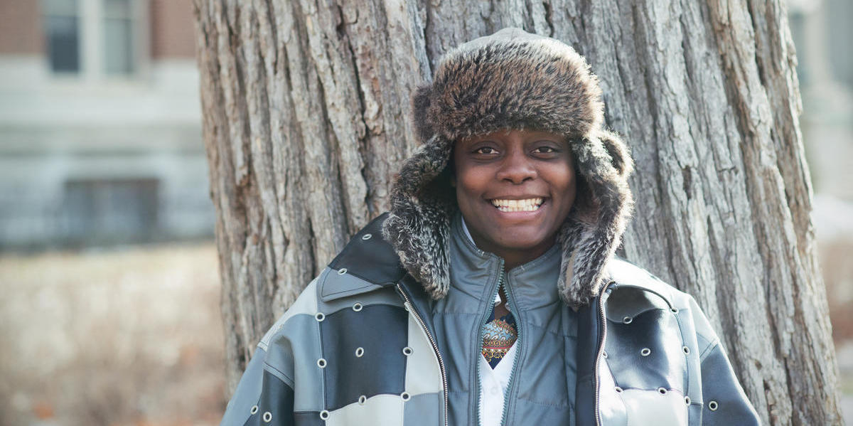 Image of someone wearing a winter jacket and hat standing against a tree and smiling.