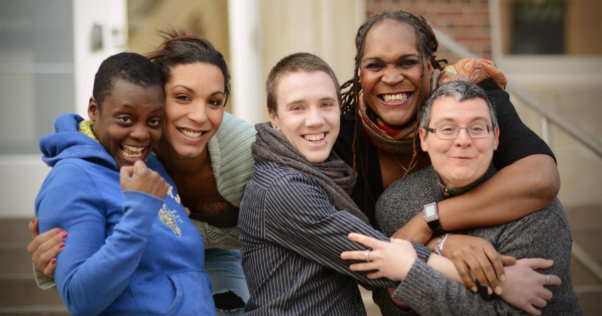 Image of five transgender individuals hugging one another while facing the camera and smiling.
